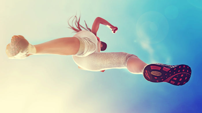 A woman in a white athletic outfit and pink-and-black running shoes is captured mid-air against a bright blue sky background.