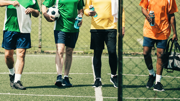 Four soccer players standing on a green turf field in front of a chain-link fence, wearing athletic jerseys in green, yellow, and orange with blue shorts. Two players hold water bottles, and one holds a soccer ball.