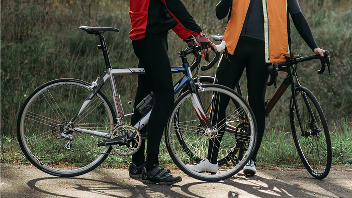 Two cyclists standing outdoors on a path with road bikes, shown from the waist down. One bike is silver and black, and the other is dark blue and black.