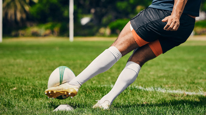 A close-up action shot of a person's legs kicking a rugby ball on a green grass field. The person is wearing black athletic shorts with orange side panels, white knee-high socks, and white cleats.