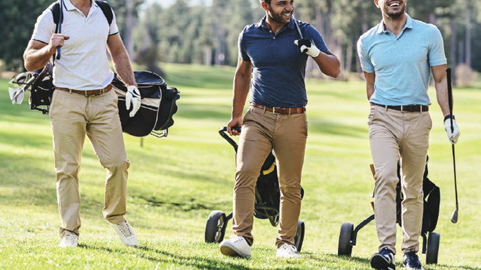Three men walking on a golf course with golf bags and a trolley, wearing polo shirts and khaki pants.