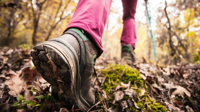 Close-up of a person hiking through a forest, showing the sole of a dark gray and black hiking boot with pink pants and a blue trekking pole in the background.