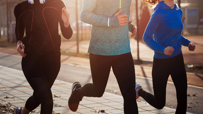 Three people jogging outdoors on a paved path during warm golden-hour sunlight. The runners are shown from the back and side, wearing athletic apparel in black, heather blue, and bright blue.