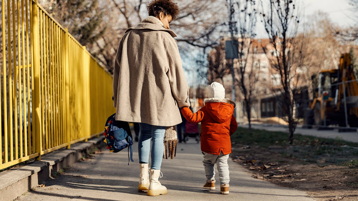 A woman and a young child walking hand in hand along a paved path outdoors, with the woman wearing a beige coat and the child wearing a bright orange jacket. A yellow fence is on the left, and trees and buildings are in the background.