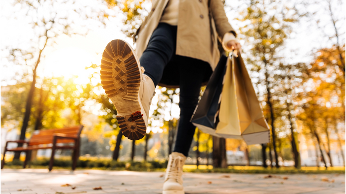 A person walking through a sunny park holding multiple shopping bags, with one boot kicked up to show the sole. The scene is backlit by warm golden sunlight with blurred autumn trees in the background.