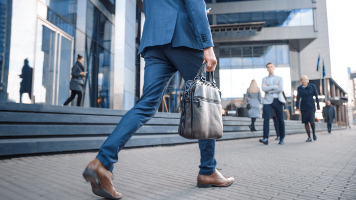 A man in a blue suit walking outdoors while carrying a gray briefcase, with blurred office building architecture and other pedestrians in the background.