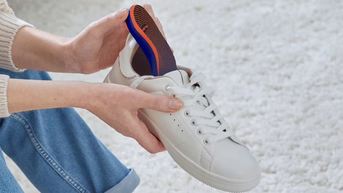 A person holding a white leather sneaker with a blue and orange insole inserted, against a soft white textured background.