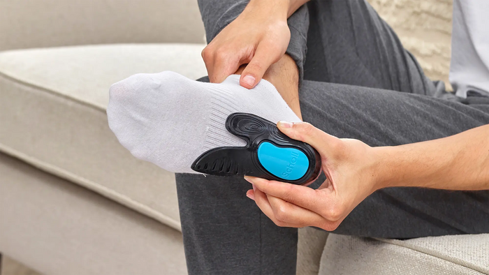 A person sitting on a sofa demonstrates a black and blue foot-shaped insert onto a white sock.