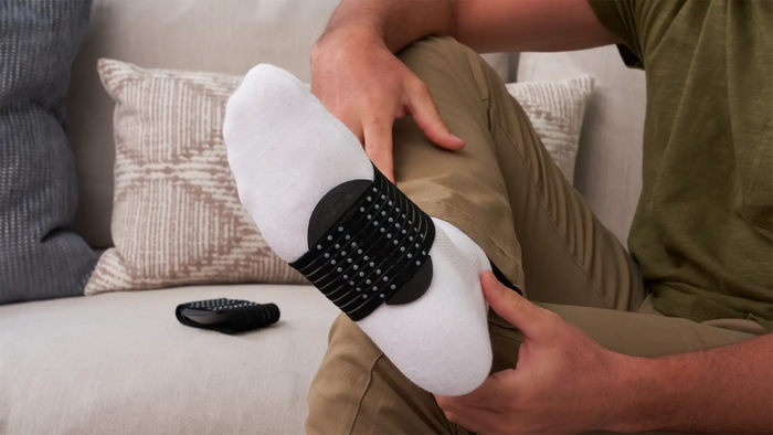 A person sitting on a couch demonstrates a white foot massager device with a black textured strap, holding the foot while a matching remote lies on the sofa.