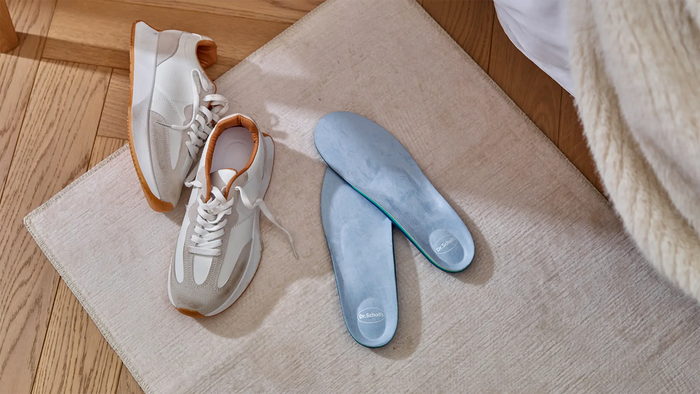 A pair of white athletic sneakers with light brown lining and gum outsoles, placed on a beige rug next to a pair of light blue insoles on a wooden floor.