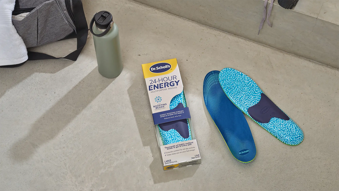 A pair of blue Dr. Scholl's 24-Hour Energy arch support insoles next to their yellow and blue retail box, with a green water bottle and a gray bag in the background on a concrete floor.