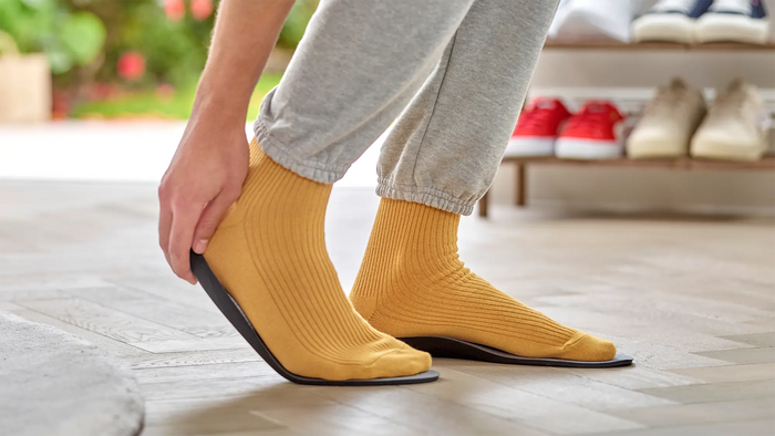 A person wearing mustard yellow ribbed slip-on socks with black wedge soles, shown from the knees down in a home setting with a shoe rack blurred in the background.