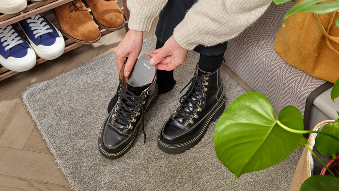 A person wearing a cream knit sweater sits on a gray rug, holding a gray foot-shaped shoe insert next to a black lace-up boot. The scene is set in a cozy room with a shoe rack, patterned rug, and a green plant in the foreground.