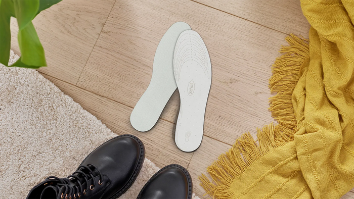 A pair of white shoe insoles placed on a light wood floor, styled with black leather lace-up boots at the bottom left and a yellow fringed throw blanket on the right.