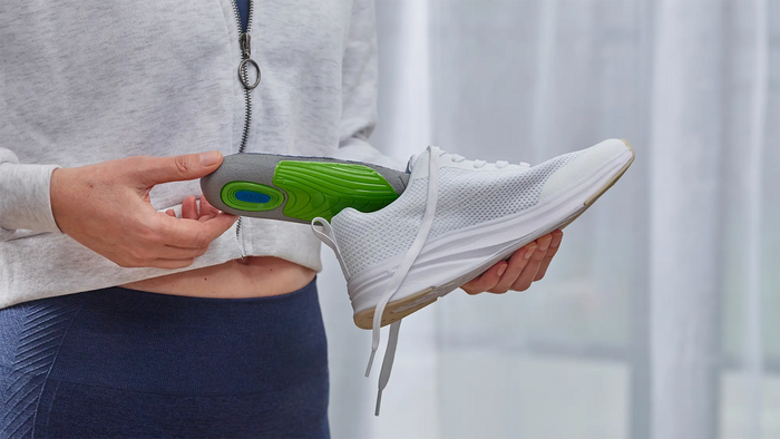 A person holding a white athletic shoe with a green and gray insole insert, shown against a soft, bright indoor background.