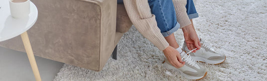 A person sitting on a beige armchair in a living room setting, tying the laces of a pair of white and gray sneakers with blue jeans and a beige knit sweater.